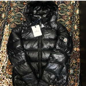 Moncler Maya  - Snygg svart jacka Moncler maya med glansig finish och klassisk logga på ärmen. Jackan har huva, dragkedja framtill och quiltad design som ger en cool och sportig vibe. Perfekt för kalla dagar när du vill hålla stilen.