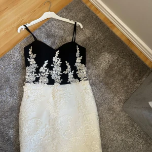 Svart och vit klänning med spetsdetaljer - Supersnygg klänning med svart överdel och smala axelband, dekorerad med vita blommiga spetsbroderier. Kjolen är vit med matchande broderade blommor och dragkedja bak. Perfekt för dig som vill sticka ut med eleganta detaljer och kontraster. Det är en 38 men passar en 36 kropp💕
