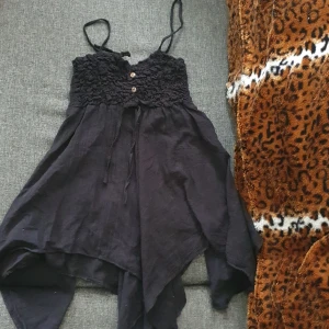 Svart kortklänning med smock och knappar - Supersöt svart kortklänning med smockad överdel, tunna axelband och dekorativa knappar framtill. Kjolen är luftig med asymmetrisk fåll och bohemisk känsla. Perfekt för varma dagar och sommarhäng.