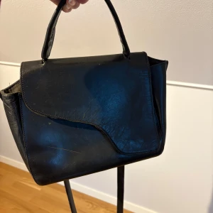 Svart handväska i skinn med axelrem från ATP atelier - Stilren svart handväska i skinn med både handtag och avtagbar axelrem. Väskan har ett unikt lock med asymmetrisk kant och silverfärgade detaljer på remmen. Perfekt storlek för det viktigaste och passar till många olika looks.