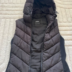 Svart dunväst med huva från Zara - Snygg svart dunväst från Zara i storlek M. Västen har quiltad design, dragkedja framtill, praktiska fickor med dragkedja och en stor huva med dragsko. Perfekt för lager-på-lager och chill höst- eller vinterstil(OOPS! ena fickans dragkedja har lite problem FINNS EN BILD😕)