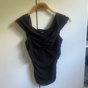 Svart omlottopp från BikBok - Svart omlottopp från BikBok med snygg drapering och twistad detalj framtill. Toppen har breda axelband och en figurnära passform som framhäver siluetten. Perfekt för dig som vill ha en stilren men ändå cool look.
