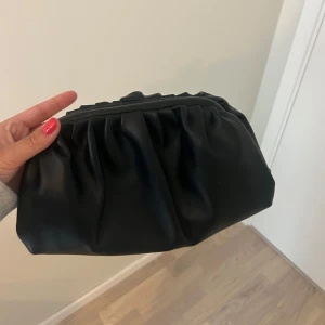 Svart kuvertväska i skinnimitation - Stilren svart kuvertväska med rynkad design och mjuk struktur. Väskan är tillverkad i skinnimitation och har en metallstängning upptill. Perfekt storlek för det viktigaste och ger en trendig touch till din outfit. Nästan aldrig använd!