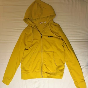 Gul hoodie från Levi's med dragkedja - Snygg gul hoodie från Levi's med svart logga på bröstet. Tröjan har huva med snörning, dragkedja framtill och två stora fickor. Perfekt för dig som gillar färg och vill ha en bekväm och stilren look.