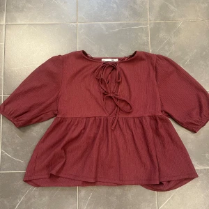 Vinröd blus med knytning från Etéa 1999 - Säljer en vinröd blus med trekvartsärm och knytning framtill från Etéa 1999. Blusen har en lös passform och är tillverkad i ett ribbat material som ger en fin struktur. Perfekt för en stilren look.