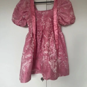 Rosa blommig klänning - Charmig rosa klänning med blommigt mönster och puffiga kortärmar. Klänningen har en fyrkantig urringning och ett band i ryggen för en söt look. Perfekt för sommarens festligheter!