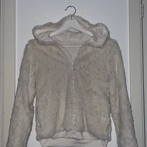 Beige teddyjacka från KappAhl - Mysig beige teddyjacka från KappAhl med dragkedja och luva. Perfekt för kyliga dagar när du vill hålla dig varm och bekväm. Jackan har långa ärmar och en mjuk, fluffig yta som ger en trendig look.