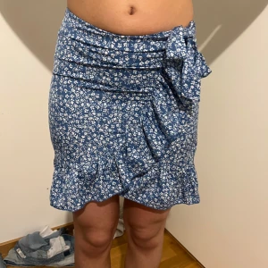 Blå blommig kjol med volang - Säljer en söt blå kortkjol med vita blommor och volangdetaljer. Kjolen har en omlott-design med knytning i midjan för en justerbar passform. Perfekt för sommarens alla tillfällen!