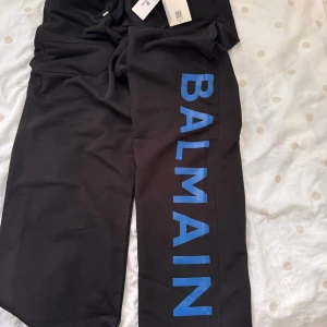 Svarta mjukisbyxor från Balmain - Snygga svarta mjukisbyxor från Balmain med blå logga längs benet. Byxorna har elastisk midja med snörning och praktiska fickor bak. Perfekta för en avslappnad stil.