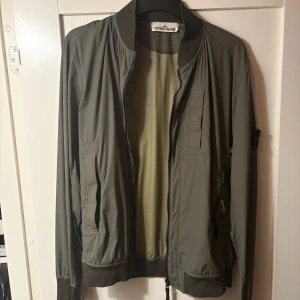 Grön bomberjacka från Stone Island - Snygg grön bomberjacka från Stone Island med dragkedja och ribbade muddar. Jackan har en klassisk design med en broderad logotyp på ärmen. Perfekt för en stilren look.