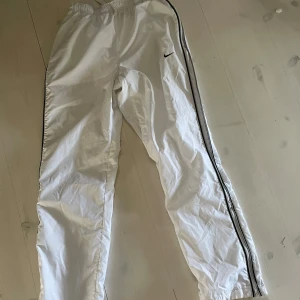 Vita träningsbyxor från Nike - Säljer ett par vita träningsbyxor från Nike med elastisk midja och svarta detaljer längs sidorna. Perfekta för sport och träning. Byxorna har en avslappnad passform och är tillverkade i ett lätt syntetmaterial.