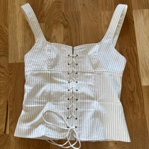Vit randig korsettopp - Snygg vit korsettopp med svarta ränder och snörning framtill. Perfekt för en stilren look med en touch av elegans. Axelbanden ger extra stöd och komfort.