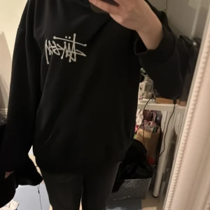Svart hoodie från Stüssy - Säljer en svart hoodie från Stüssy med deras ikoniska logga tryckt i vitt på bröstet och på ryggen. Hoodien har en klassisk passform med långa ärmar och en bekväm huva. Perfekt för en avslappnad stil.
