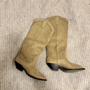 Beige cowboy boots i mocka - Snygga beige cowboy boots i mocka med spetsig tå och dekorativa sömmar. Perfekta för en stilren look med en touch av vilda västern. De har en låg klack och är knähöga, vilket ger en klassisk cowboy-stil. Köpta 500 kr