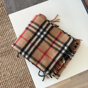 Burberry Beige rutig halsduk - Säljer en klassisk beige halsduk med rutigt mönster i svart, rött och vitt. Den har fransar i ändarna och är perfekt för att hålla dig varm under kyliga dagar. Passar både till vardags och mer uppklädda tillfällen.