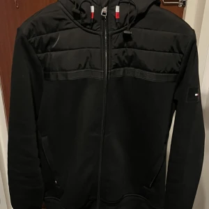Svart jacka med huva - Snygg svart jacka med huva och dragkedja framtill. Jackan har quiltade detaljer på axlarna och en liten logotyp på ärmen. Perfekt för kyligare dagar.