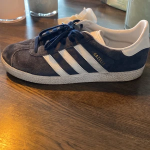 Blå Adidas Gazelle sneakers - Snygga blå Adidas Gazelle sneakers med vita ränder och snörning. Skorna har en klassisk design med en ovandel i mocka. Perfekta för en stilren look. Pris kan diskuteras 