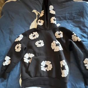 Denim tears hoodie - Säljer en cool svart hoodie från denim tears med vita blommor över hela tröjan och huvan. Den har en praktisk magficka och är perfekt för en avslappnad stil. Passar bra för både vardag och chill.