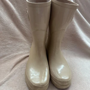 Beige stövlar med spänne - Snygga beige stövlar i syntetmaterial med dekorativt spänne på sidan. Perfekta för regniga dagar och ger en stilren look. De har en klassisk design som passar till många olika outfits.