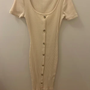 Beige ribbad klänning med knappar - Säljer en stilren beige ribbad klänning med knappar framtill. Klänningen har korta ärmar och en rund halsringning. Ny!!