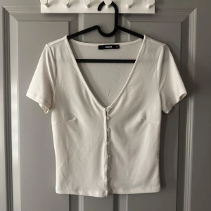 Vit kortärmad topp från Bik Bok - Säljer en stilren vit topp från Bik Bok med korta ärmar och v-ringning. Toppen har knappar framtill och är gjord i ett mjukt material. Finns en liten fläck men syns inte så bra (se bild)