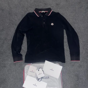 Svart pikétröja från Moncler - Snygg svart pikétröja från Moncler som är i storlek S och riktigt fint skick! Tröjan är självklart äkta. Moncler tillbehör ingår i köpet. Har ni frågor är det bara kontakta!