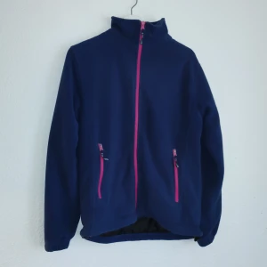 Mörkblå fleecejacka från Stormberg - Säljer en mörkblå fleecejacka från Stormberg med rosa dragkedjor och broderad logga på ärmen. Jackan har en hög krage och två fickor med dragkedja framtill. Perfekt för kyliga dagar!