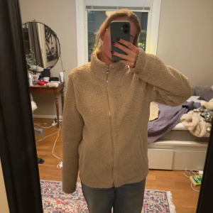 Beige teddyjacka från H&M - Mysig beige teddyjacka från H&M med dragkedja framtill. Jackan har en normal passform och är perfekt för kyligare dagar men passar ändå som kofta till sommarkvällar. Den har långa ärmar och en hög krage för extra värme. Perfekt för att hålla stilen och värmen samtidigt!