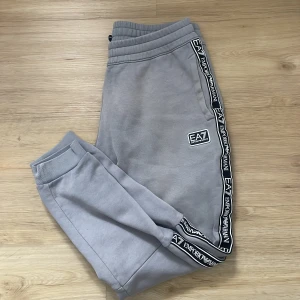 Grå mjukisbyxor från Emporio Armani - Snygga grå mjukisbyxor från Emporio Armani med EA7-logga på sidan och längs benen. Byxorna har en elastisk midja och muddar vid bensluten för en bekväm passform. Perfekta för en avslappnad stil.