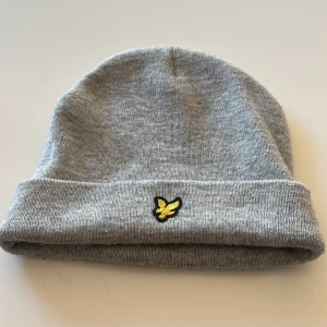 Grå mössa från Lyle & Scott - Snygg grå mössa från Lyle & Scott med ett broderat gult emblem framtill. Perfekt för kyliga dagar med sin mjuka och bekväma passform.