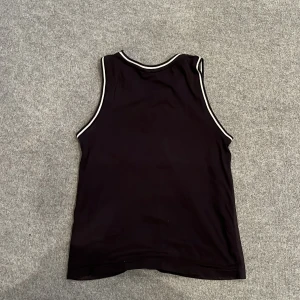 Svart ärmlös basketlinne - Snygg svart ärmlös basketlinne med vit kant och tryck av en basketspelare och korg. Perfekt för sportiga aktiviteter och träning. Stort nummertryck på framsidan.