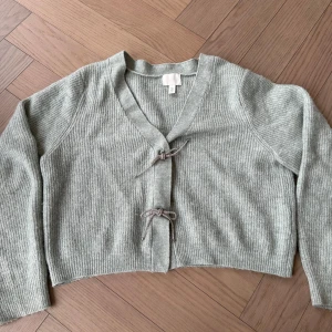 Grön ribbad kofta från H&M - Säljer en stilren grön ribbad kofta från H&M. Koftan har en v-ringad design med knytning framtill och långa ärmar. Passar både till jeans och kjol för en avslappnad look. Knapp stängs med knäppning 