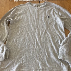 Grå långärmad tröja från Ralph Lauren - Säljer en stilren grå långärmad tröja från Ralph Lauren med den klassiska loggan broderad på bröstet. Perfekt för en avslappnad och snygg look.