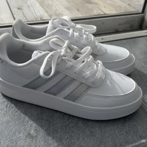 Vita sneakers från Adidas - Nypris: 900kr skick: aldrig använda. Pris går att förhandla. Snygga vita sneakers från Adidas med klassiska tre ränder i silver på sidan. Skorna har snörning och en stilren design som passar perfekt till vardags. Tillverkade i syntetmaterial för en modern look. 