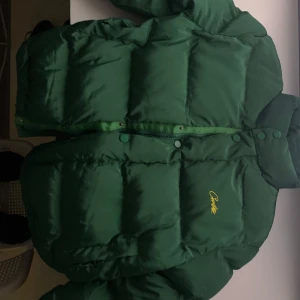 Grön pufferjacka från Corteiz - Säljer en snygg grön pufferjacka från Corteiz med gul insida. Jackan har en dragkedja och knappar framtill samt en broderad logga på bröstet. Perfekt för kyliga dagar!