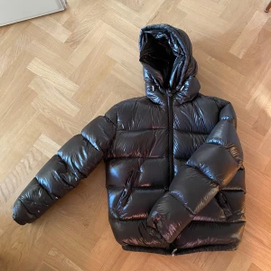 Olivgrön pufferjacka från Zara - Säljer en mörkgrön pufferjacka från Zara med huva och dragkedja. Jackan är perfekt för kyliga dagar och har en glansig finish som ger en modern look. Den är vadderad och har långa ärmar för extra värme.