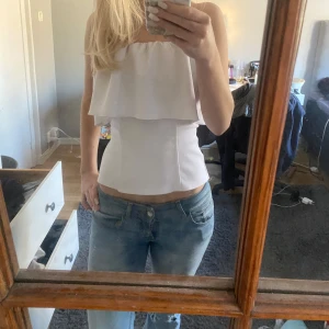 Vit axelbandslös topp - Säljer en stilren vit axelbandslös topp med volangdetalj. Perfekt för sommarens/höst/vårens varma dagar eller en kväll ute. Toppen har en tight passform och är tillverkad i ett mjukt material.