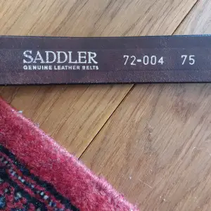 Snyggt brunt läderbälte från Saddler med en klassisk design.  Det svarta bältet har en robust metallspänne och är märkt med 'RAGE for Leather'. Tillverkat i Sverige med respekt för människa och natur.Perfekt för att komplettera din outfit.