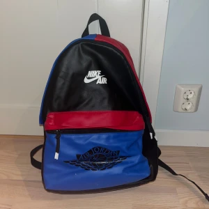 Nike air jordan ryggsäck - Snygg ryggsäck från Nike Air i svart, röd och blå färgkombination. Väskan har en dragkedja och ett stort fack med Air Jordan-logga på framsidan. Perfekt för att bära med sig allt man behöver under dagen.