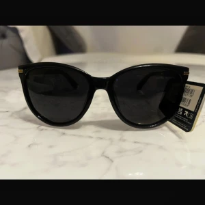 Svarta solglasögon från Foster Grant - Snygga svarta solglasögon från Foster Grant med polariserade linser som minskar bländning och ger 100% UV-skydd. De har en klassisk design med en lätt böjd form och subtila gulddetaljer vid gångjärnen.