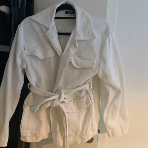 Vit sommar/ vår jacka med bälte - Snygg vit sommar/vår jacka från Gina Tricot   med knytbälte i midjan. Jackan har långa ärmar och två bröstfickor med lock. Perfekt för en stilren look, knappt använd därav säljer. 