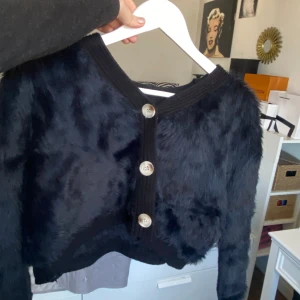 Svart pälsliknande kofta med knappar - Säljer en svart kofta med pälsliknande textur och stora knappar framtill. Koftan har en croppad passform och långa ärmar, perfekt för att ge en trendig touch till din outfit.