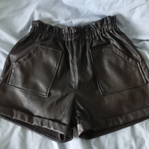 Svarta shorts i syntetmaterial - Snygga svarta shorts med elastisk midja och stora framfickor. Perfekta för en cool och avslappnad stil. De har en glansig finish som ger en modern touch.