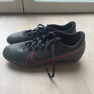 Svarta fotbollsskor från Nike - Säljer ett par svarta fotbollsskor från Nike med röda detaljer och snörning. Skorna har en klassisk design med dobbar för bra grepp på planen. Perfekta för fotbollsträning och matcher. Skorna är relativt använda därav lite smutsiga men funkar precis som de ska och funkar jätte bra till fotboll. Storlek 39 men skulle säga närmare 38 då de är väldigt små i storlek.