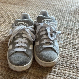 Grå Adidas Campus sneakers mocka - Säljer ett par grå Adidas Campus sneakers i mjuk mocka med vita klassiska ränder på sidorna och vit sula. Skorna har rund tå, platt sula och vita snören. Inte så jätte använda men växte ur de ganska fort. 