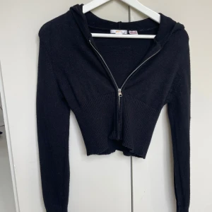 Svart croppad hoodie från Gina Tricot - Svart croppad hoodie från Gina Tricot med ribbad midja och långa ärmar. Tröjan har huva och dragkedja framtill, vilket gör den både snygg och chill. Materialet är stickat och känns mjukt mot huden. Perfekt för dig som gillar en trendig och bekväm stil.