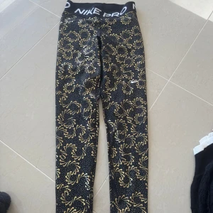 Nike Pro Dri-Fit leggings med guldmönster - Nike Pro Dri-Fit leggings i svart med coolt guldigt blommönster och små prickar. Hög midja med bred resår och Nike Pro-logga. Superskön stretchig syntet som andas, perfekt för träning eller gym. Snygg passform som sitter tight och följer kroppen.