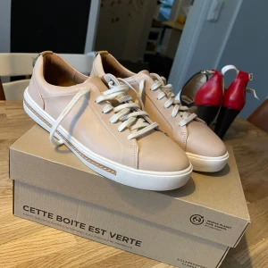 Beige sneakers från Clarks - Snygga beige sneakers från Clarks med vit platt sula och rund tå. Skorna har vita snören och är tillverkade i slätt skinn. Stilren design med diskreta sömmar och neutrala färger som passar till många outfits.Helt nya,puderrosa 
