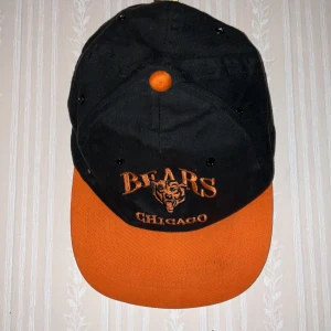 Chicago Bears keps svart och orange - Snygg keps från Chicago Bears med broderad logga och text i orange på svart tyg. Skärmen och toppen har stark orange färg, resten är svart. Klassisk form med ventilationshål och justerbar passform. Materialet är 100% bomull, perfekt för sport och streetwear.
