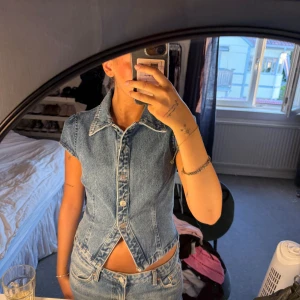 Jeansblus med krage från Peppermayo - Säljer en snygg jeansblus från Peppermayo i klassisk blå denim. Västen har korta ärmar, markerad krage och knappar framtill. Figurnära passform med coolt omlottliknande snitt nedtill! Aldrig använt! nypris 865kr